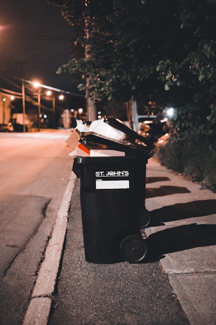 A black wheeled rubbish bin placed on a residential sidewalk at night, with the label 'ST. JOHN'S' on the front. The bin is overflowing with various discarded items including brown and white cardboard