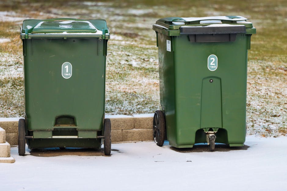 A pile of assorted household rubbish and waste items scattered on the ground amidst dry grass and twigs. The collection includes cardboard boxes, plastic containers in various colors, a small pink pla